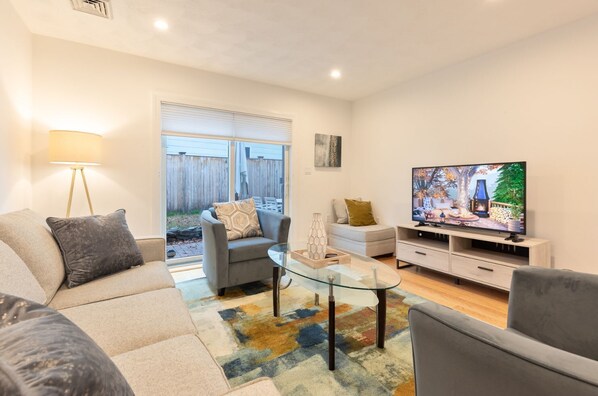 A modern living room with a sofa, armchair, glass coffee table, TV, and a view of the backyard.