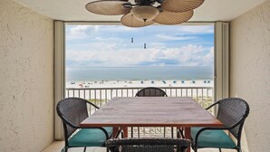 Ein Balkon mit Holztisch und Korbstühlen mit Blick auf einen Strand, auf dem Menschen und Sonnenschirme sind.