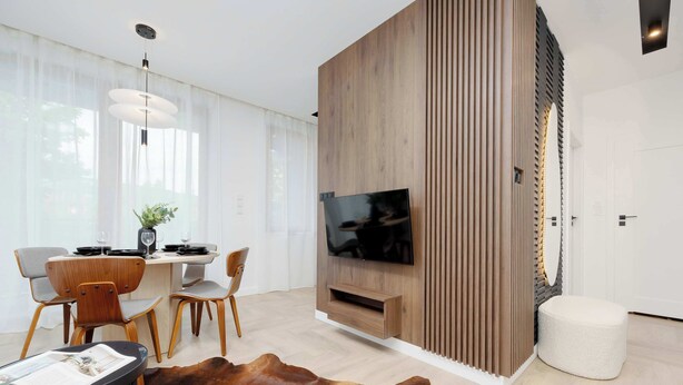 Een moderne woonkamer met een flatscreen televisie aan een houten wand, een eethoek met een tafel en stoelen, en een groot leren vloerkleed.