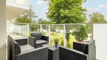Ein Balkon mit Korbmöbeln und Glasgeländer, der auf eine grüne Rasenfläche und Bäume blickt.