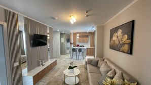 Ein modernes Wohnzimmer mit einer Couch, einem Couchtisch und einem an der Wand montierten Fernseher. Im Hintergrund befindet sich eine Küche mit Barhocker und einem Esstisch.