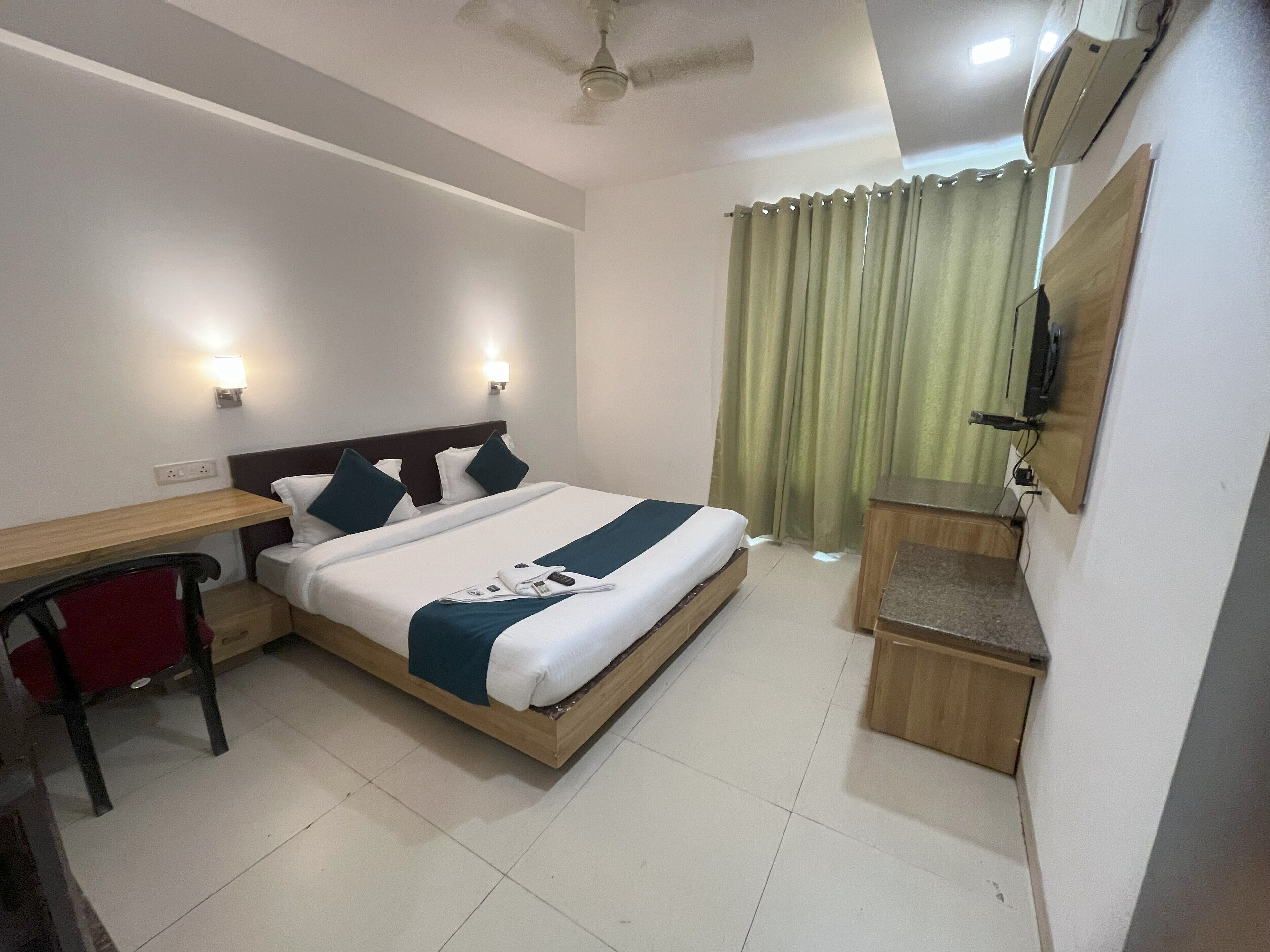 A hotel room with a bed, bedside table, chair, desk, and a TV mounted on the wall.