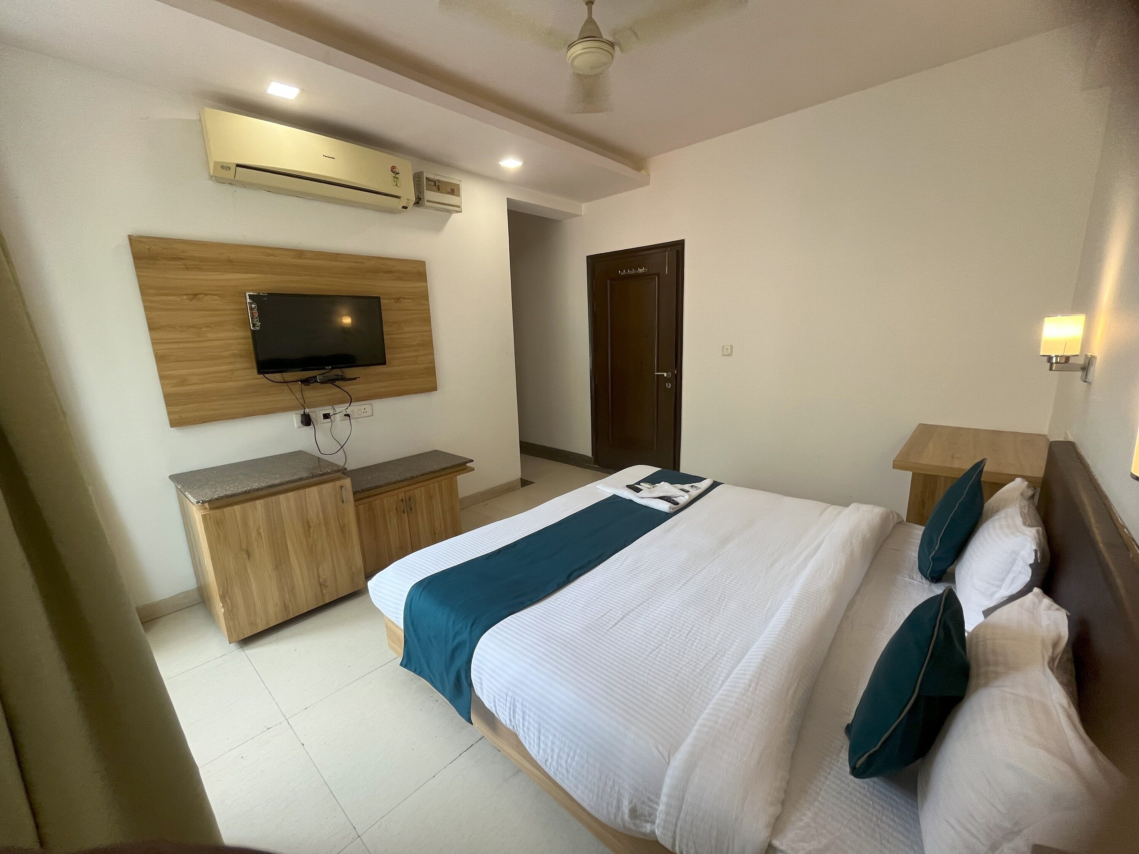 A hotel room with a bed, a TV mounted on a wooden panel, a desk with a chair, and an air conditioning unit.