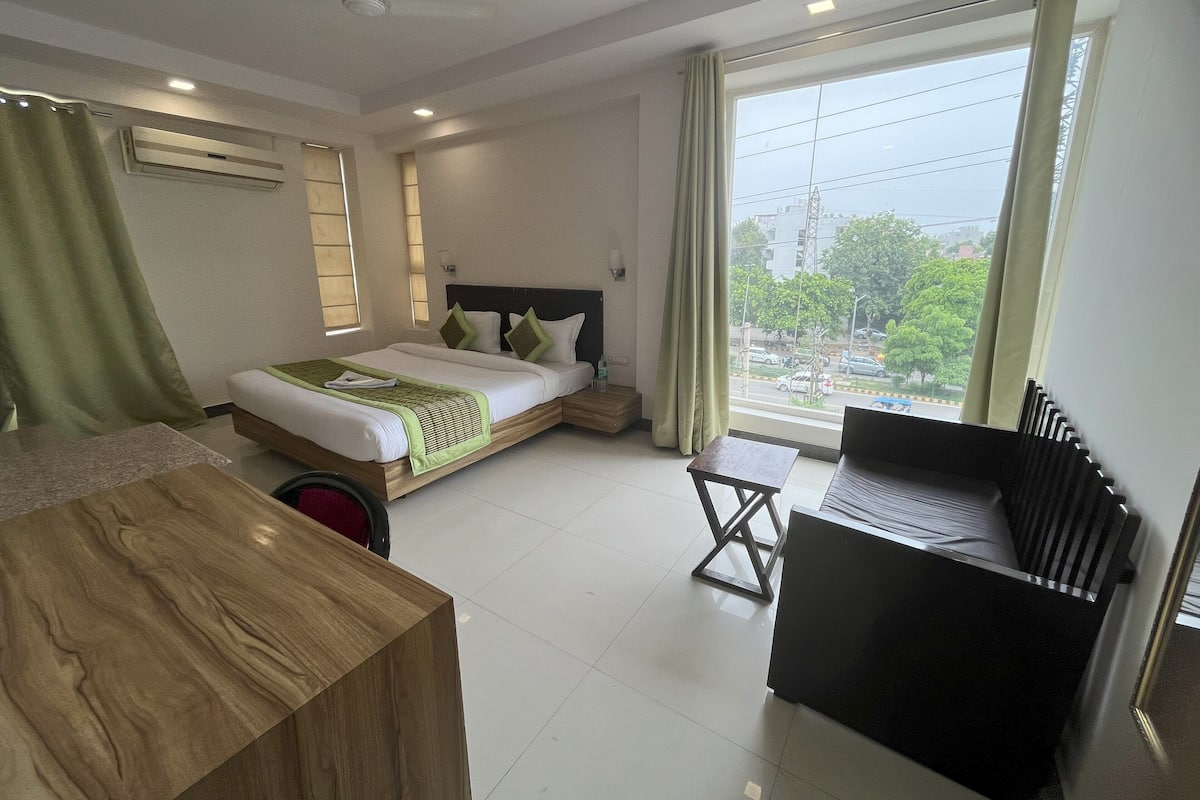 A hotel room with a bed, a desk, a chair, a window with a view of the city, and an air conditioning unit.