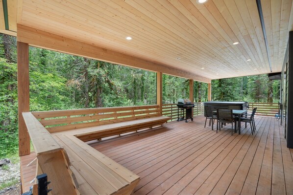 Une terrasse en bois avec un banc et un espace barbecue, le tout entouré d’une forêt dense.