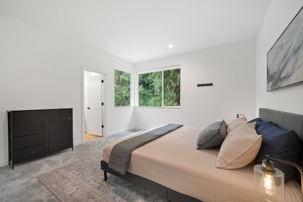 Un dormitorio con cama, cómoda, ventana con vista al bosque y un cuadro en la pared.
