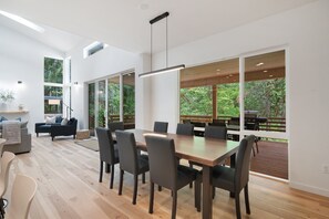 Un vaste espace à manger avec une grande table et des chaises en bois, de grandes fenêtres donnant sur une terrasse et la verdure, et un luminaire moderne au plafond.