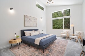 A modern bedroom with a large bed, a side table, a chair, a floor lamp, and a large window with a view of trees. - Sandy River Group Getaway 3 (Mount Hood Village)