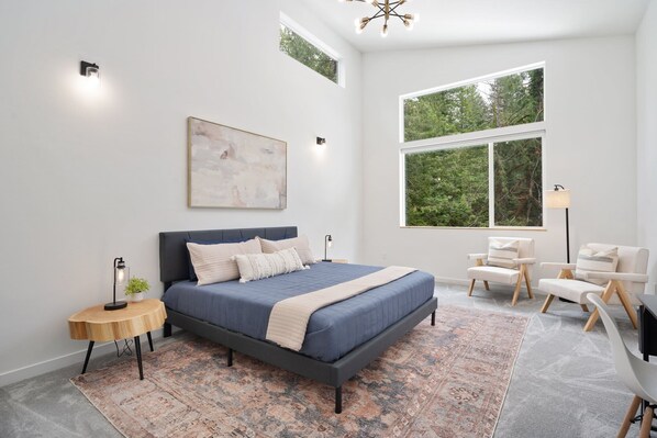 A modern bedroom with a large bed, a side table, a chair, a floor lamp, and a large window with a view of trees.