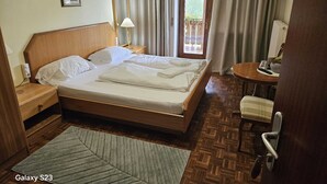 A hotel room with a wooden bed, a bedside table, a lamp, a small table with a chair, and a view of the outdoors through a door. - Etwas Älter Aber Fein (Villnöss)