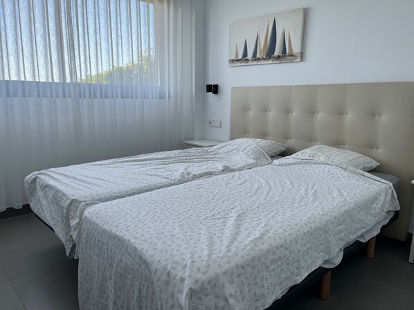 A bedroom with two beds, a window with sheer curtains, a painting of sailboats, and a light-colored headboard. - Apartment - Sleeps 4 - Parking - Garden - Pool (Alhaurin el Grande)