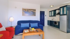 A living room with a blue sofa, a wooden coffee table with a fruit basket, a remote control, and a book. There is a kitchen area with a refrigerator, microwave, and cabinets. - Immaculate 2-bed Apartment No 5 in Playa Blanca (Yaiza)