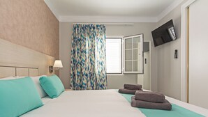Una habitación de hotel con una cama, un televisor montado en la pared, una ventana con cortinas y un perchero.