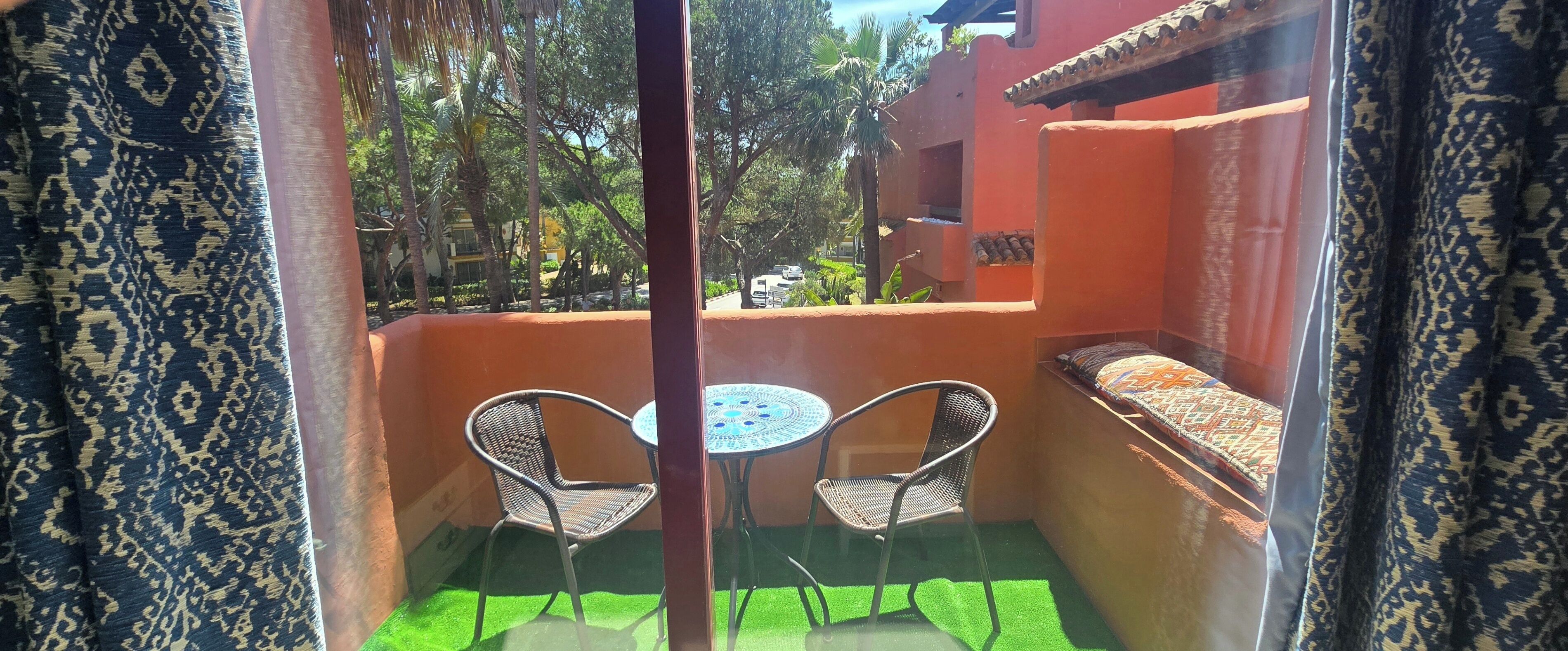 A balcony with two chairs, a small table, and a view of a building with a red exterior.