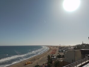 Ein Strand mit Sonnenschirmen und Menschen, Gebäude im Hintergrund und ein klarer Himmel.