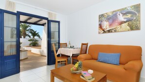 A living room with a sofa, a coffee table with fruits and books, a dining area with a table and chairs, and a large window with a view of palm trees. - Impeccable 2-bed Apartment No 2 in Playa Blanca (Yaiza)