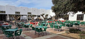 An outdoor dining area with multiple tables and chairs, a building with 'Spanglish' signs, and a large tree providing shade. - Stylish 2-bed Apartment Overlooking the Fairways (Torre-Pacheco)