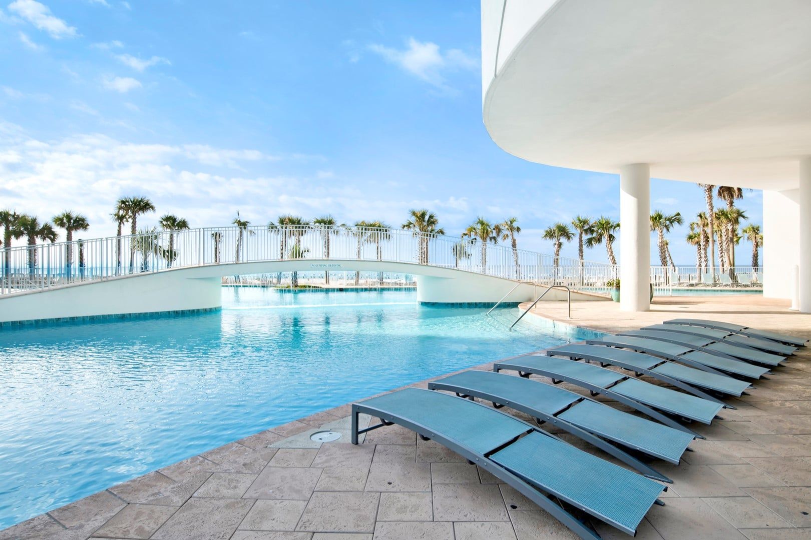 A swimming pool with a bridge, palm trees, and lounge chairs.