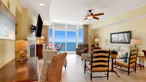 A spacious living room with a large window offering a view of the ocean, a ceiling fan, and a variety of seating options including wicker chairs and a sofa. - TURQUOISE Place.  3 bed 3 bath.  Sleeps 12. Balcony Hot Tub.  August 25% off. (Orange Beach)