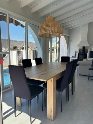 A dining area with a wooden table and black chairs, large windows with a view of the sea, and a hanging wicker light fixture. - Casa Amira a Modern House With Pool (Pulpi)