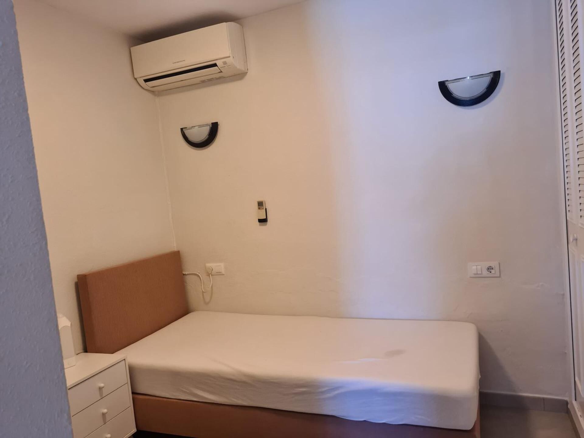 A small, single-bed room with a brown headboard, white bedding, an air conditioning unit mounted on the wall, and a small bedside table with drawers.