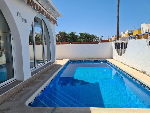 A swimming pool with clear blue water, surrounded by a white wall and a wooden deck. - Casa Amira a Modern House With Pool (Pulpi)
