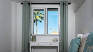 Un dormitorio con cama, escritorio con cajones, ventana con cortinas y vista a palmeras y un edificio.