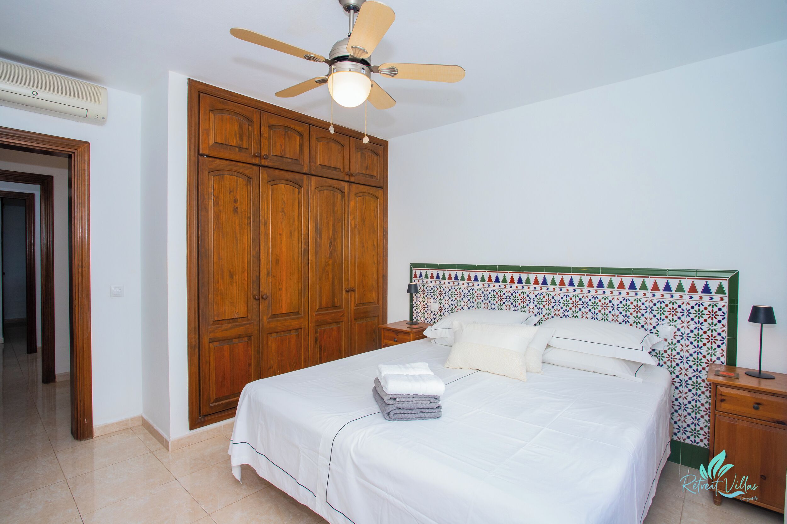 A bedroom with a bed, a ceiling fan, wooden wardrobe, and a tiled wall.