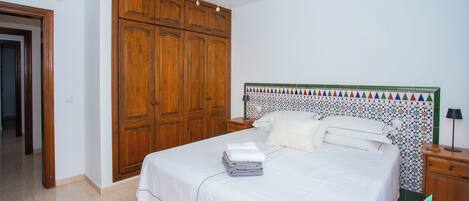 A bedroom with a bed, a ceiling fan, wooden wardrobe, and a tiled wall.