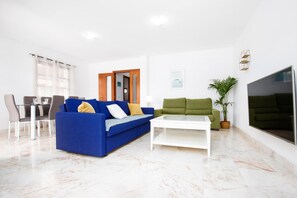 Un salon avec un canapé bleu, une table basse blanche, un canapé vert, un coin repas avec des chaises et un grand téléviseur fixé au mur.