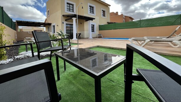 A patio with a glass table and chairs, a pool, and a house with a tiled roof and columns. - House - Sleeps 6 - Pool - Pet Friendly (La Oliva)