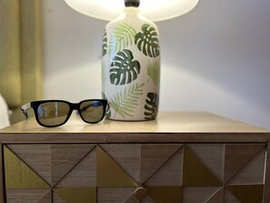 Un par de gafas de sol sobre una mesita de noche de madera con un diseño geométrico, junto a una lámpara con motivos vegetales.