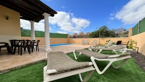 A poolside area with lounge chairs and a table, a pergola, and a view of a swimming pool and distant buildings. - House - Sleeps 6 - Pool - Pet Friendly (La Oliva)