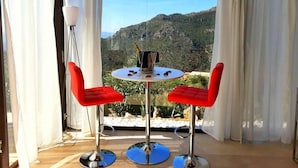 En balkon med et bord og to røde stole, en flaske champagne og udsigt til et bjergrigt landskab.