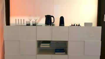 Uma unidade de estantes moderna, branca e modular com um tampo de vidro. Contém uma cafeteira, um bule e algumas chávenas. Também há dois livros na prateleira inferior.
