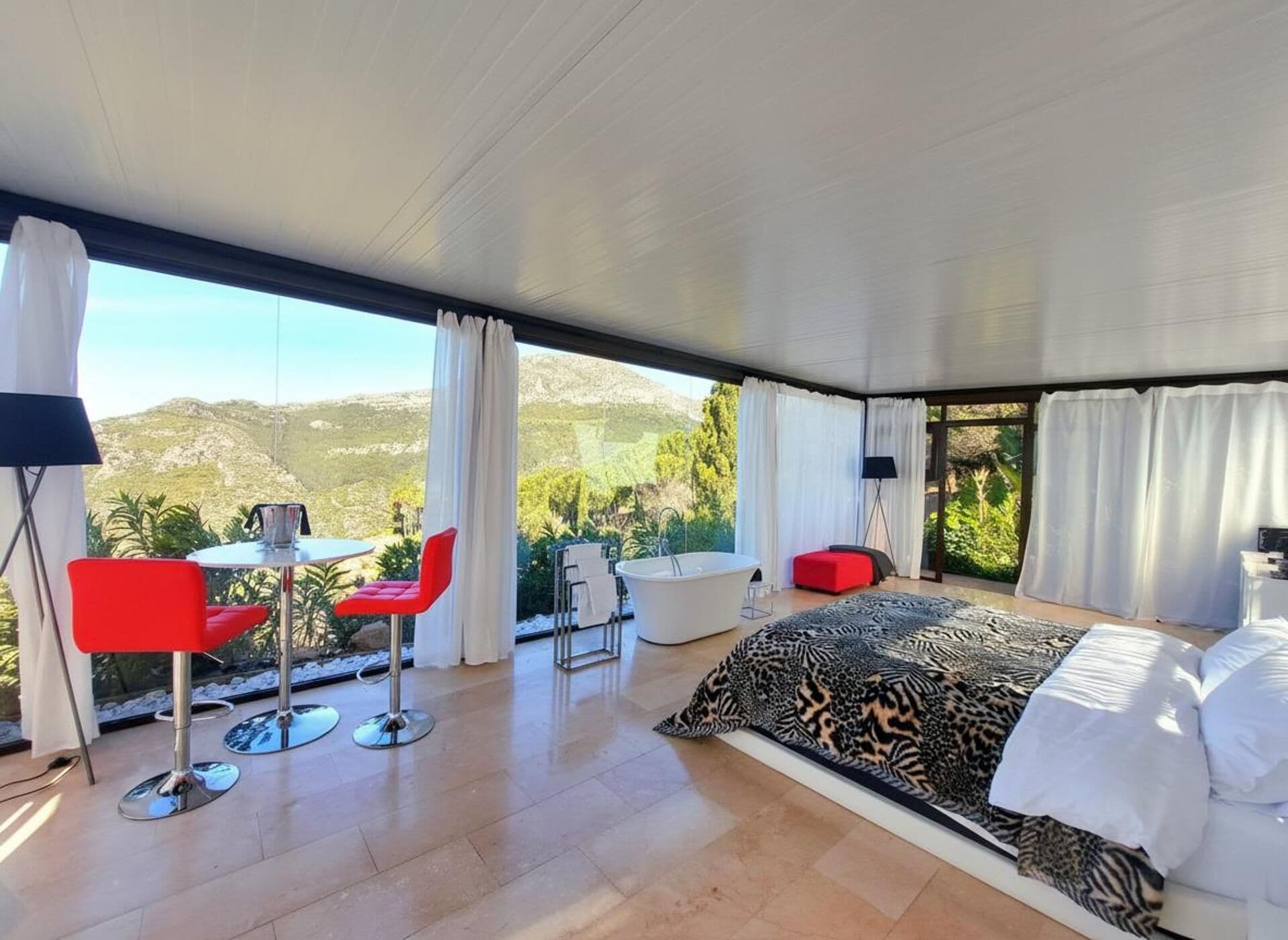 A modern bedroom with a large bed, a sitting area with red chairs, a bathtub, and a view of the mountains.