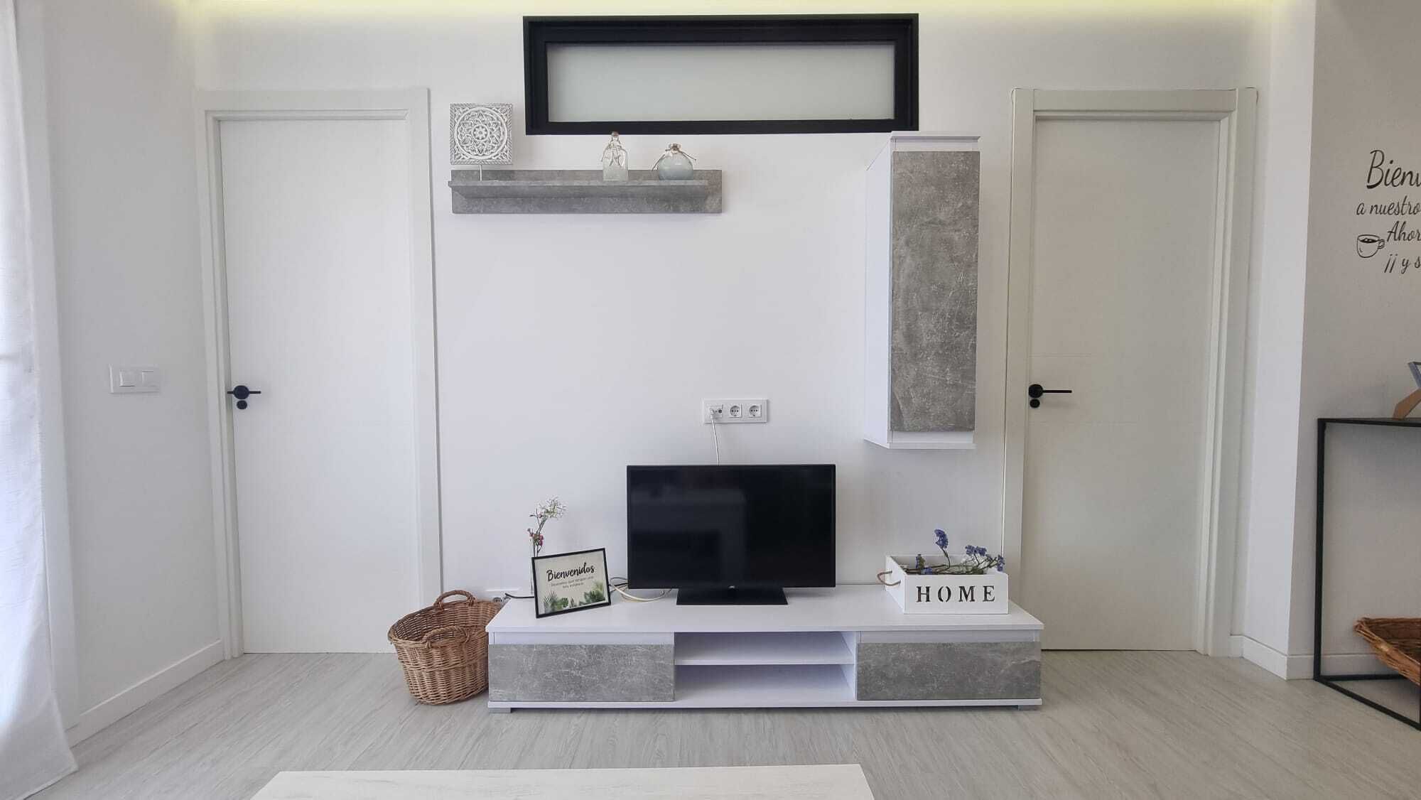 Una sala de estar moderna con un televisor de pantalla plana sobre un mueble gris, una pieza decorativa en la pared encima del televisor, y un letrero que dice 