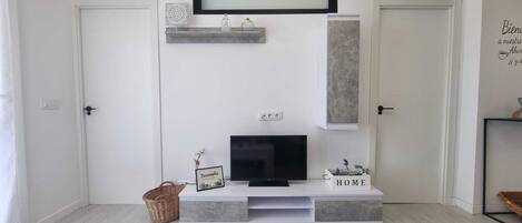Una sala de estar moderna con un televisor de pantalla plana sobre un mueble gris, una pieza decorativa en la pared encima del televisor, y un letrero que dice