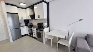 Een kleine keuken met een koelkast, fornuis, magnetron en koffiezetapparaat. Ook aanwezig: een wasmachine, een eettafel met stoelen en een vloerlamp.
