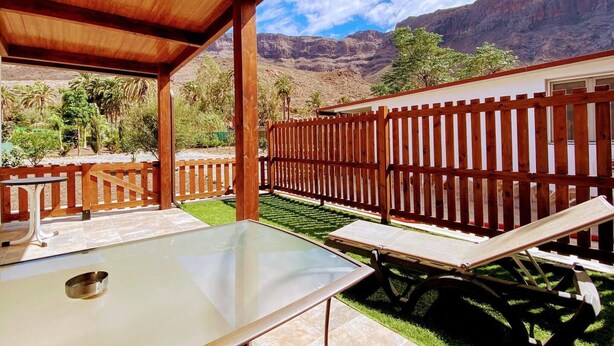 Un patio couvert avec une table en verre et deux fauteuils de jardin, une clôture en bois et une vue sur les montagnes.