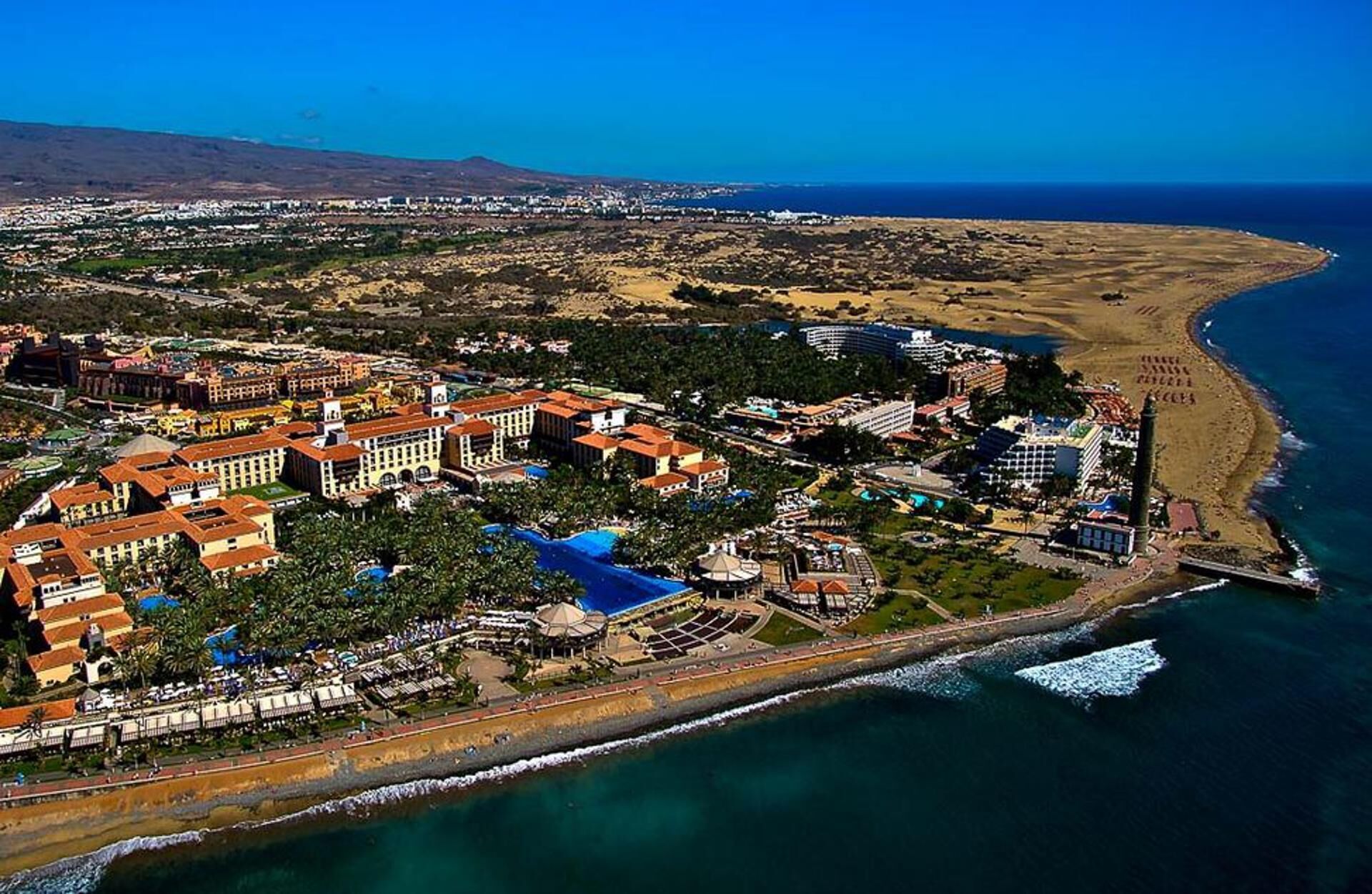 Vista aérea de un complejo turístico costero con varios edificios, una playa y un muelle.