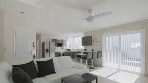 Un salón moderno con un sofá blanco, cojines negros, una mesa de centro de vidrio y una cocina al fondo.