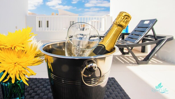 Eine Flasche Champagner in einem Metallkübel mit einem Glas, gelben Blumen und einem Liegestuhl auf einem Balkon mit einem weißen Gebäude und blauem Himmel im Hintergrund.