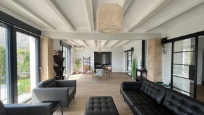 A spacious living area with a large black leather sofa, a matching ottoman, and a dining table with chairs. There is a large window providing a view of the outdoors, and a hanging light fixture above the seating area. - Modern 5 Bed Villa in Javea (Javea)