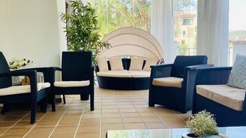 Una habitación iluminada por el sol con muebles de mimbre, una planta grande en maceta y vistas de edificios a través de una ventana.