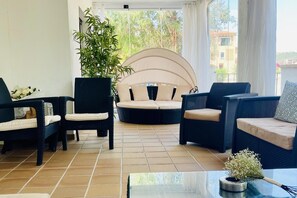 A sunlit room with wicker furniture, a large potted plant, and a view of buildings through a window. - 2 Bed Flat-sleeps 4-pets-parking-marina Duquesa (Manilva)
