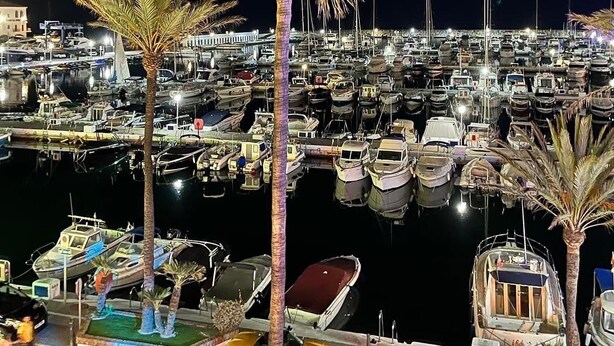 Un port de plaisance de nuit, avec de nombreux bateaux amarrés, des palmiers et une zone riveraine bien éclairée.