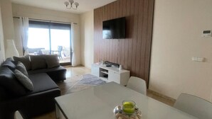 Un salon moderne avec un canapé, un téléviseur fixé sur un mur recouvert de panneaux en bois, une table à manger avec une bougie, et une vue sur l’extérieur grâce à une grande fenêtre.