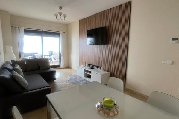 Ein modernes Wohnzimmer mit einer Couch, einem an einer Holzpaneelwand montierten Fernseher, einem Esstisch mit Kerze und Blick ins Freie durch ein großes Fenster.