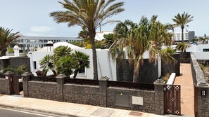 Un edificio moderno con un muro de piedra, palmeras y una entrada protegida por una reja.